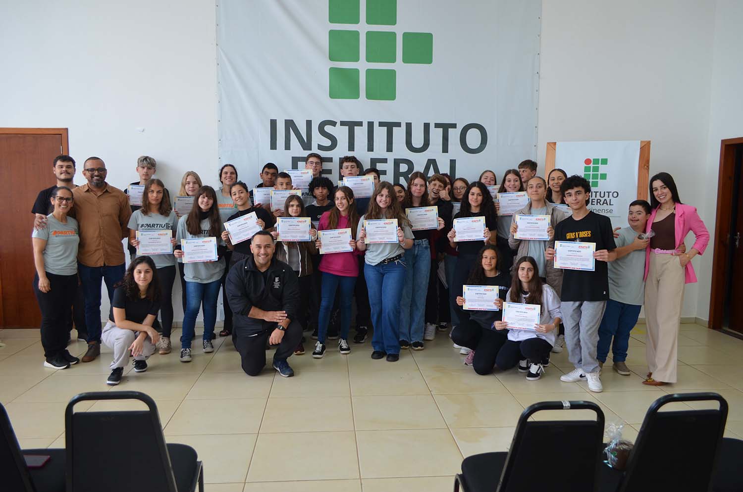 A imagem mostra um ambiente iluminado, com piso claro e cadeiras pretas aparecendo parcialmente no primeiro plano. Há um grupo de estudantes, professores, coordenadores e monitores posando para uma foto coletiva em frente a um painel com a logo do IFC Concórdia. Os estudantes têm certificados nas mãos e a maioria das pessoas está sorrindo para a câmera.