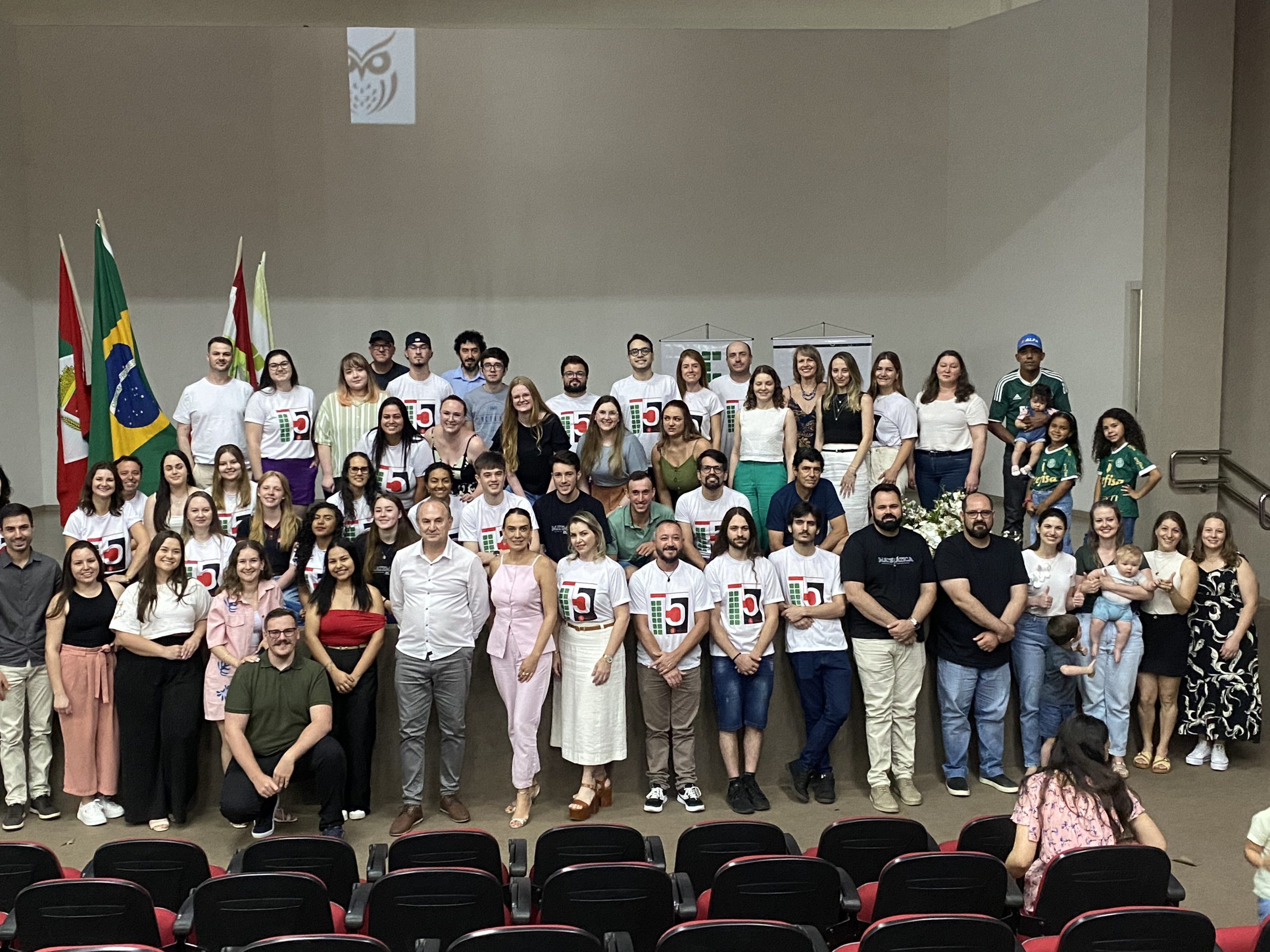 Foto colorida, em ambiente interno, registrada em no palco de um auditório. No centro da imagem há aproximadamente cinquenta pessoas organizadas em várias fileiras. A maioria está em pé; algumas pessoas estão agachadas ou sentadas na parte frontal. Grande parte do grupo veste camisetas brancas com a estampa do número “15”, enquanto outras pessoas usam roupas variadas, em cores claras e escuras. Há diversidade de gêneros, idades, tons de pele e estilos. Ao fundo, à esquerda, estão duas bandeiras hasteadas: a do Brasil e a de Santa Catarina. Em ambos os lados do palco, há arranjos florais brancos e verdes em vasos metálicos. As pessoas sorriem e olham em direção à câmera.
