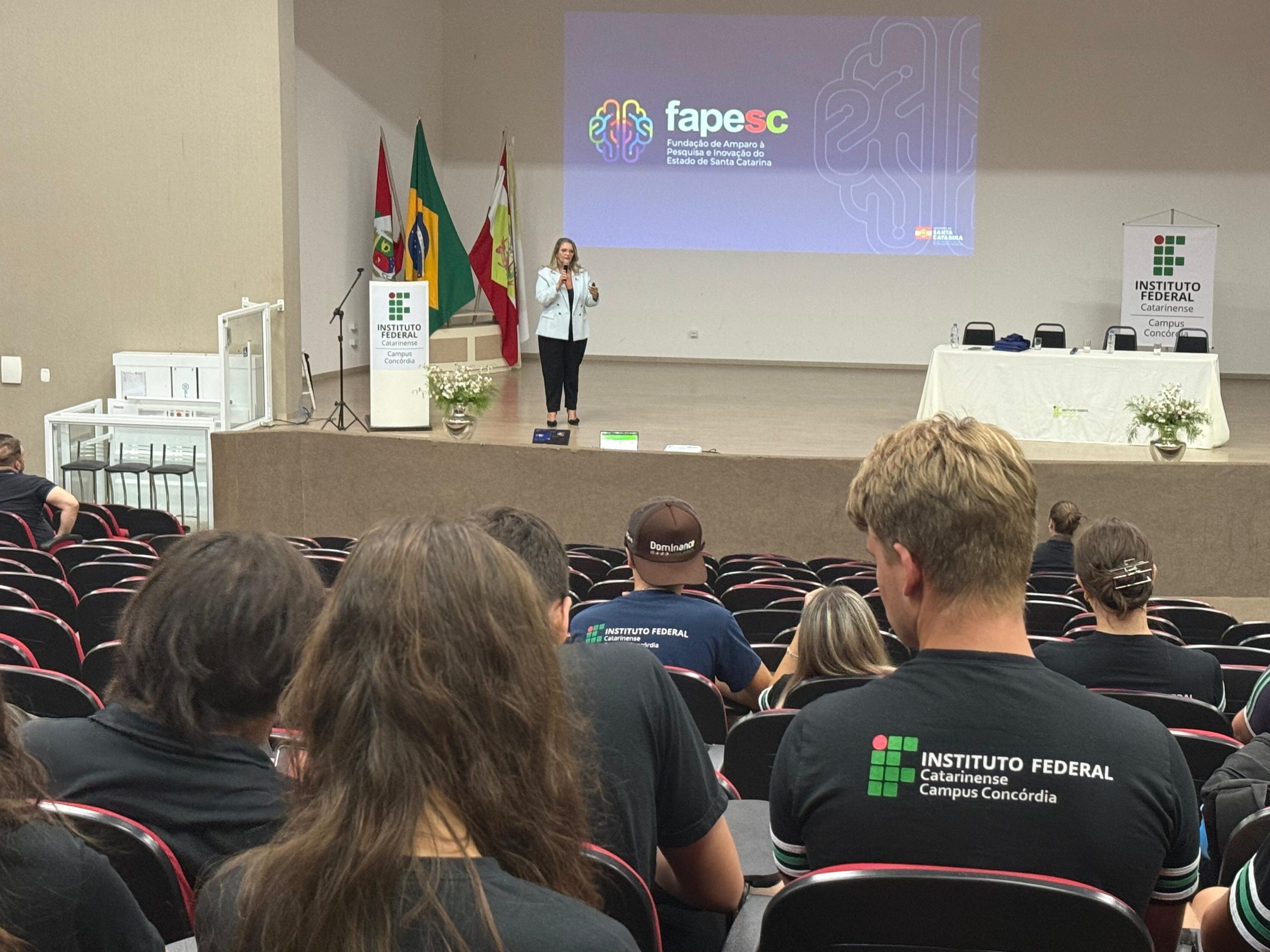 Foto colorida de um auditório durante uma palestra. Em primeiro plano, aparecem pessoas sentadas em cadeiras pretas com detalhes vermelhos, vistas de costas. Algumas usam camisetas pretas com a identificação do “IFC Concórdia”. No palco, ao centro, uma mulher (Valeska Daniela Tratsk) está em pé segurando um microfone e falando ao público. Ela veste blazer claro, blusa escura e calça preta. Ao fundo, há uma projeção em tela grande com o logotipo da FAPESC e o texto “Fundação de Amparo à Pesquisa e Inovação do Estado de Santa Catarina”. À esquerda do palco, há um púlpito com a marca do Instituto Federal Catarinense e três bandeiras posicionadas atrás. À direita, uma mesa com toalha branca, cadeiras e arranjos de flores. O ambiente é amplo, com paredes claras e iluminação interna.