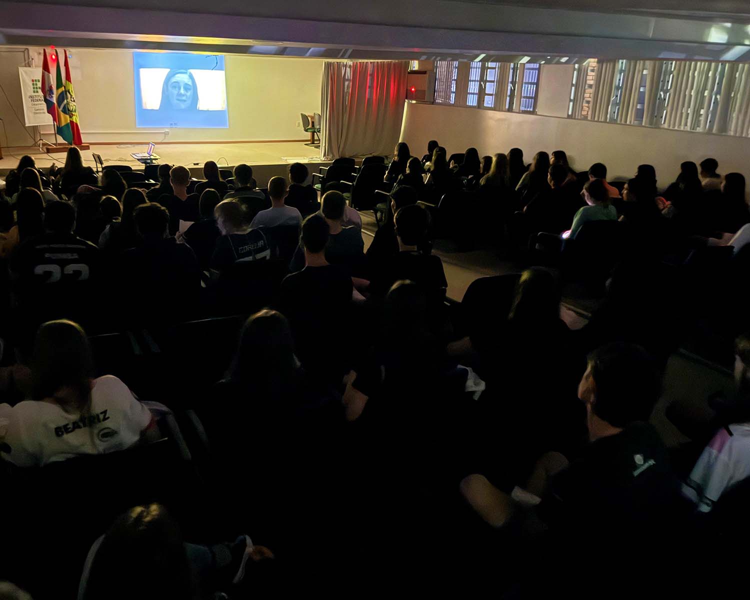 Imagem colorida de ambiente interno, um auditório. O ambiente está na penumbra. Os estudantes estão assistindo à exibição de um filme projetado à frente. Na tela aparece o rosto de uma mulher.