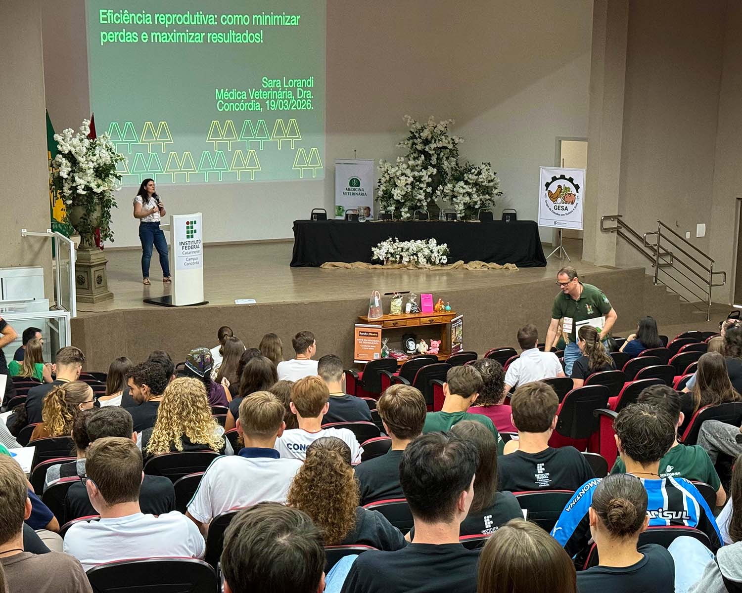 Auditório com público sentado assistindo a uma apresentação. No palco, uma mulher fala ao microfone ao lado de um púlpito com o logotipo do Instituto Federal. O slide projetado exibe o título “Eficiência reprodutiva: como minimizar perdas e maximizar resultados!”, com nome da palestrante e data. Há bandeiras, arranjos de flores e banners institucionais no palco.