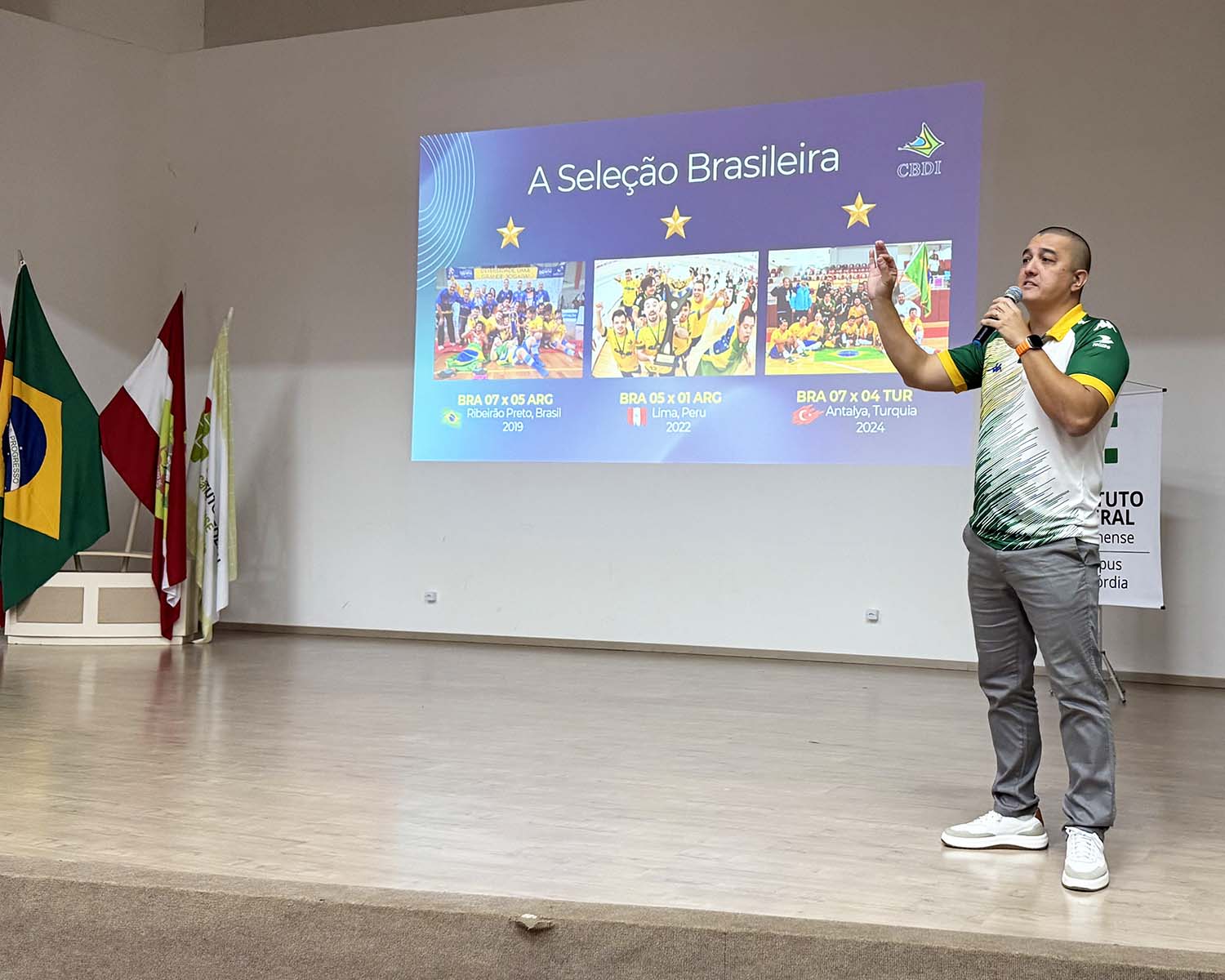 Homem em pé (Alessandro Cardoso) sobre um palco segura um microfone e gesticula com a outra mão durante uma apresentação. Ele veste camiseta esportiva nas cores verde, amarelo e branco, calça cinza e tênis branco. Ao fundo, um slide projetado traz o título “A Seleção Brasileira” e mostra fotos e resultados de jogos. À esquerda do palco, há bandeiras; à direita, aparece parcialmente um banner do Instituto Federal.