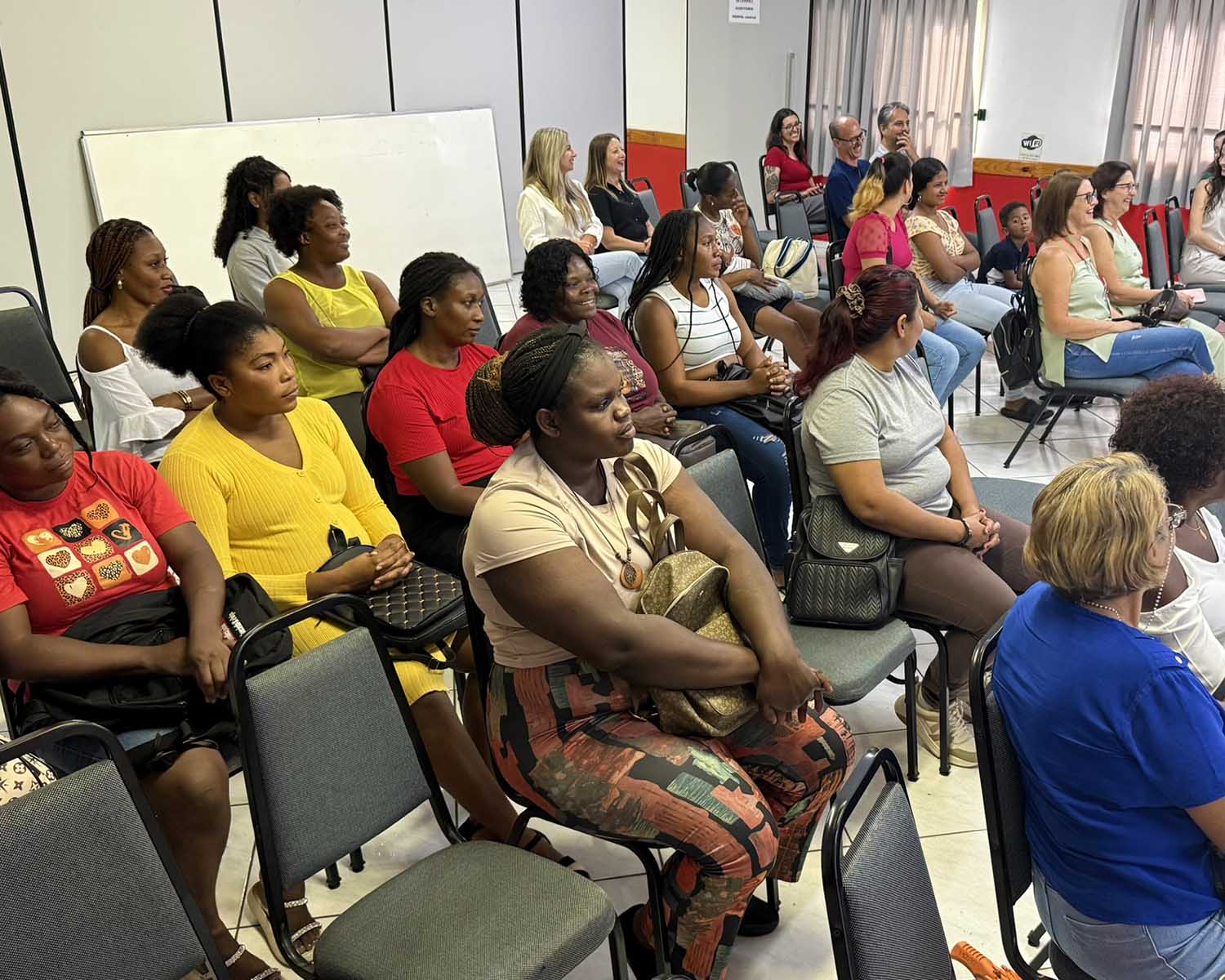 Sala com cerca de 20 pessoas sentadas em cadeiras voltadas para um palco, durante a aula inaugural do curso. A maioria é de mulheres negras, usando roupas casuais coloridas. Algumas sorriem e observam atentamente alguém fora do enquadramento. Ao fundo, há outras participantes, incluindo mulheres brancas, homens e uma criança. O ambiente é iluminado, com paredes claras e cortinas nas janelas.